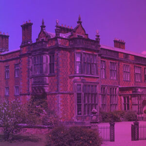A large, old brick mansion with intricate detailing and tall chimneys, bathed in a gradient of purple and pink light, looks as if it’s illuminated by distant fireworks.