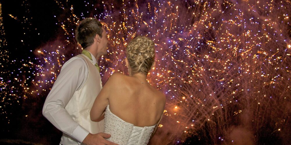 A bride and groom facing a sky filled with brilliant fireworks, with the groom's hand gently resting on the bride’s waist.