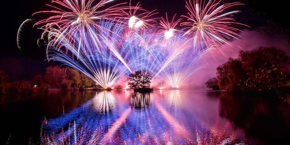 060A6590 (1) Colorful fireworks bursting over a serene lake, illuminating the trees and their reflections in the water.